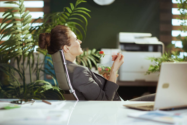 Sustainable workplace. relaxed modern female employee in a grey business suit in modern green office with laptop eating salad. - Фото, изображение