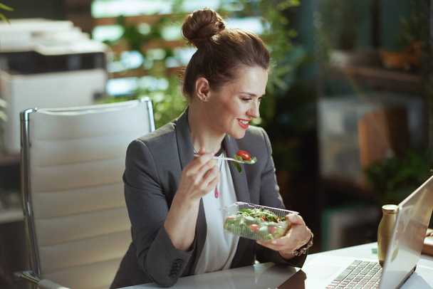 Sustainable workplace. happy modern middle aged accountant woman in a grey business suit in modern green office with laptop eating salad. - Φωτογραφία, εικόνα