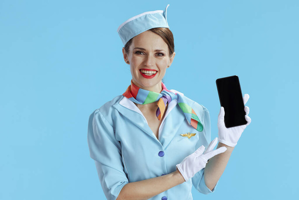 happy modern air hostess woman isolated on blue background in blue uniform showing smartphone blank screen. - Фото, зображення