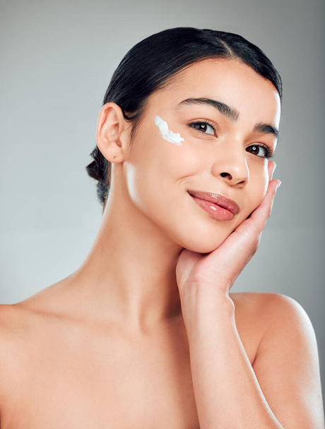 Studio Portrait of a beautiful smiling mixed race woman applying cream to her face. Hispanic model with glowing skin grey copyspace background. - Foto, Imagem