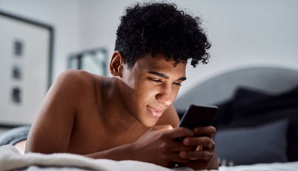 Поддерживать социальную связь во времена социального дистанцирования. молодой человек, использующий смартфон, расслабляясь дома на кровати - Фото, изображение