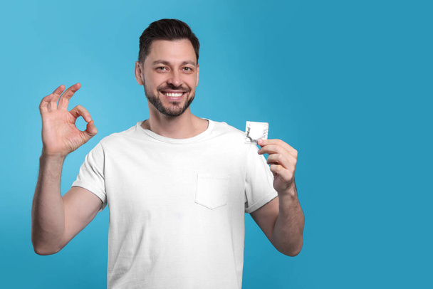 Happy man with condom showing ok gesture on light blue background. Space for text - Fotografie, Obrázek