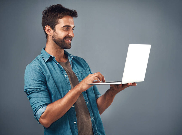 Blogcuların en iyi arkadaşı. Yakışıklı bir gencin gri bir arkaplanda dizüstü bilgisayar kullanırken çekilmiş bir fotoğrafı. - Fotoğraf, Görsel