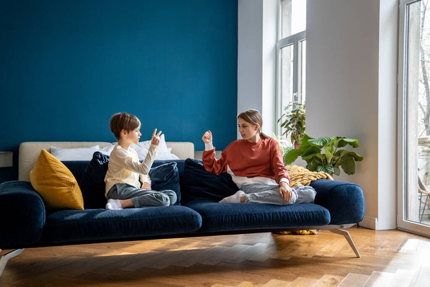 Children siblings sitting on sofa playing fun kids game Rock Paper Scissors to determine who has to clean room, brother and sister spending time together at home, family leisure concept - Valokuva, kuva