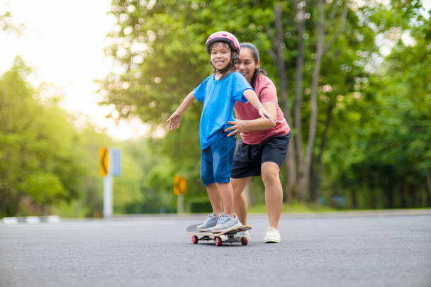 Active little girl and mom enjoy skateboarding. Cute little girl wearing helmet practicing skateboarding in park. Mother trains her daughter to skateboard. Outdoor sports for children. - Фото, изображение