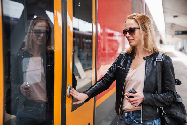 Νεαρή ξανθιά γυναίκα με τζιν, πουκάμισο και δερμάτινο μπουφάν που φοράει τσάντα και γυαλιά ηλίου, πατάει το κουμπί πόρτα του σύγχρονου τρένου ταχύτητας για να επιβιβαστεί στο σταθμό του τρένου. Ταξίδια και μεταφορές. - Φωτογραφία, εικόνα