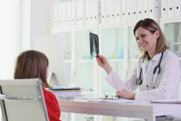 Γυναίκα παιδίατρος με χαμόγελο κρατά X-ray του μικρού ασθενούς κάθεται στο τραπέζι στο χώρο εργασίας. Schoolgirl εξετάζει το γιατρό και ακτινογραφία εικόνα - Φωτογραφία, εικόνα