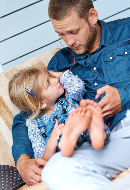 What should we play next. an adorable little girl sitting on the sofa sharing headphones with her father - Foto, Bild