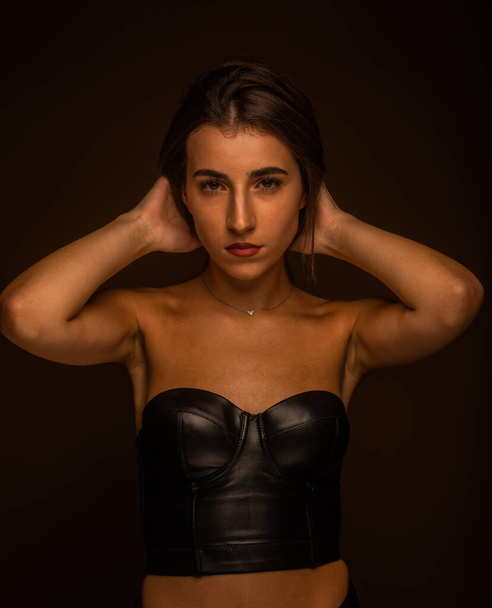 A sexy brunette girl in black leather top fixing her hair while posing in studio. Seductive looking. Close up photo - Photo, image