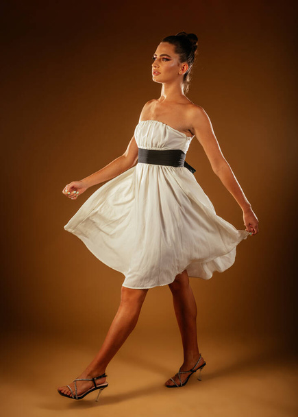 Sexy girl in white outfit and high heels walking in studio on a light brown background. Side view - Fotó, kép