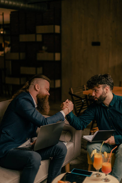 Young bearded serious businessman with his laptop in his lap making a deal with the other business man and sealing the deal with a firm handshake - Photo, image