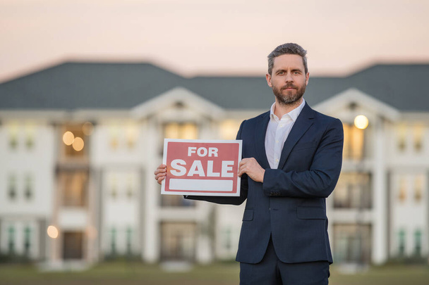 Successful real estate agent in suit with sign for sale. Real estate broker front of new house. Real estate home owner business man salesman sell house, new apartment in modern building - 写真・画像