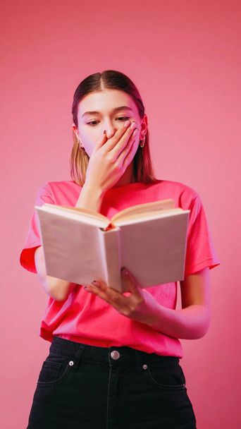 Překvapivé čtení. Šokovaná literatura. Ohromený vzrušený dívka drží knihu pokrývající ústa strach s storyline román izolované na neonové barvy růžové pozadí. - Fotografie, Obrázek