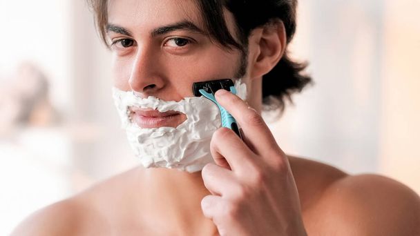 Shaving procedure. Facial grooming. Male skincare. Attractive undressed man with foam on face using razor morning routine in light room. - 写真・画像