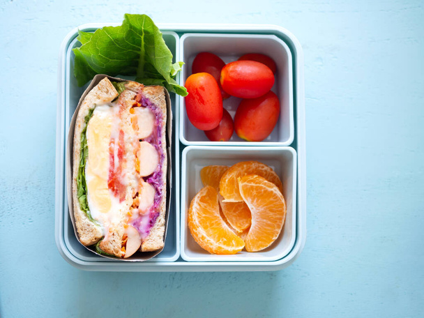 Σπιτικό υγιεινό μπλε κουτί φαγητού με σάντουιτς και πορτοκάλι, λαχανικά ντομάτας, σε μπλε ξύλινο φόντο. Κάτοψη, με χώρο αντιγραφής. - Φωτογραφία, εικόνα