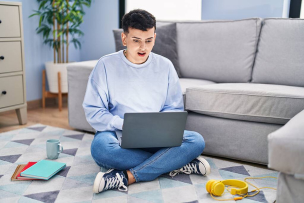Non binary person studying using computer laptop sitting on the floor afraid and shocked with surprise expression, fear and excited face.  - Foto, Imagem