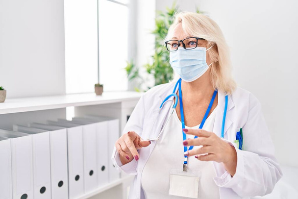 Middelbare leeftijd blonde vrouw dragen arts uniform en medisch masker spreken in de kliniek - Foto, afbeelding