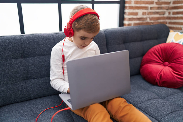 Adorable garçon hispanique utilisant ordinateur portable et écouteurs assis sur le canapé à la maison - Photo, image