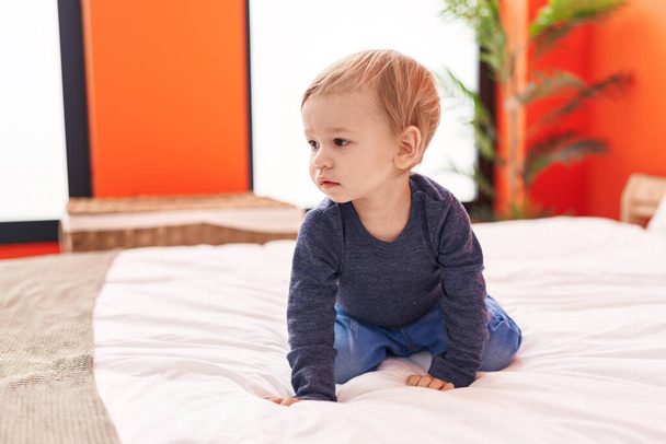 Liebenswertes blondes Kleinkind sitzt mit entspanntem Gesichtsausdruck im Schlafzimmer auf dem Bett - Foto, Bild