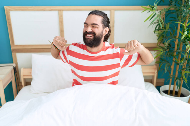 Young hispanic man waking up stretching arms at bedroom - Photo, Image
