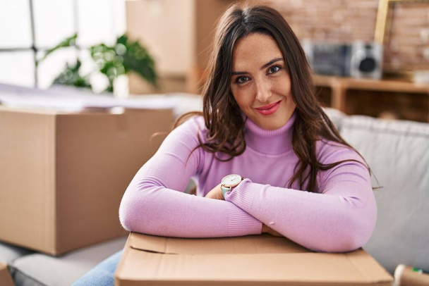 jonge latino vrouw glimlachen zelfverzekerd leunend op pakket in nieuw huis - Foto, afbeelding