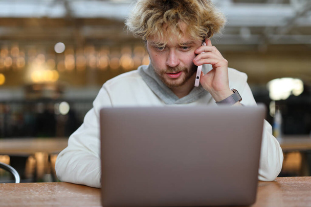 Concentré jeune homme aux cheveux bouclés sur le lieu de travail parlant au téléphone derrière un ordinateur portable. - Photo, image