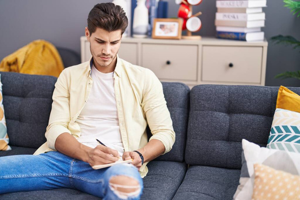 Fiatal spanyol férfi ír jegyzetfüzetbe ül a kanapén otthon - Fotó, kép