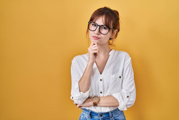 Young beautiful woman wearing casual shirt over yellow background looking confident at the camera smiling with crossed arms and hand raised on chin. thinking positive.  - Photo, Image