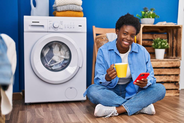 Afro-Amerikaanse vrouw met behulp van smartphone en het drinken van koffie wachten op wasmachine in de wasruimte - Foto, afbeelding