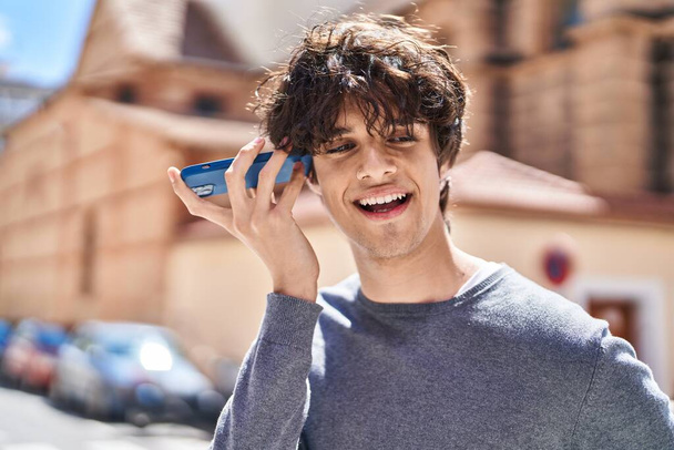 Joven hombre hispano sonriendo confiado escuchando mensaje de audio por el teléfono inteligente en la calle - Foto, imagen