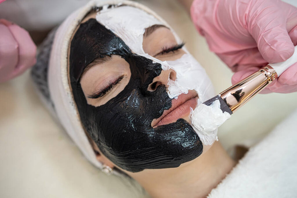 Schöne junge Frau erhalten weiße und schwarze Gesichtsmaske auf der Hälfte des Gesichts auf dem Wellnessbereich liegend. Hautpflegekonzept - Foto, Bild