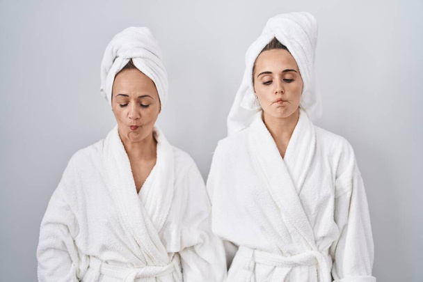 Middle age woman and daughter wearing white bathrobe and towel making fish face with lips, crazy and comical gesture. funny expression.  - Фото, изображение