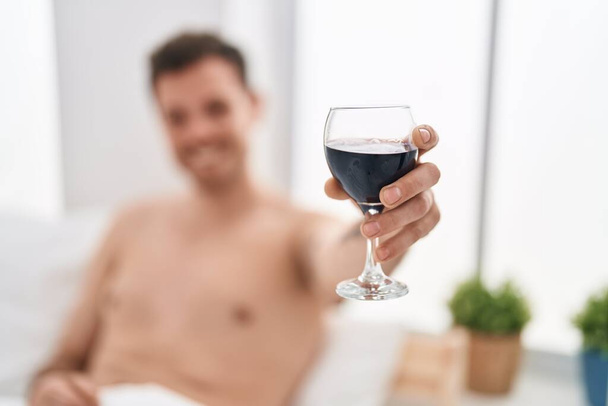 Junger hispanischer Mann trinkt Wein auf Bett im Schlafzimmer - Foto, Bild