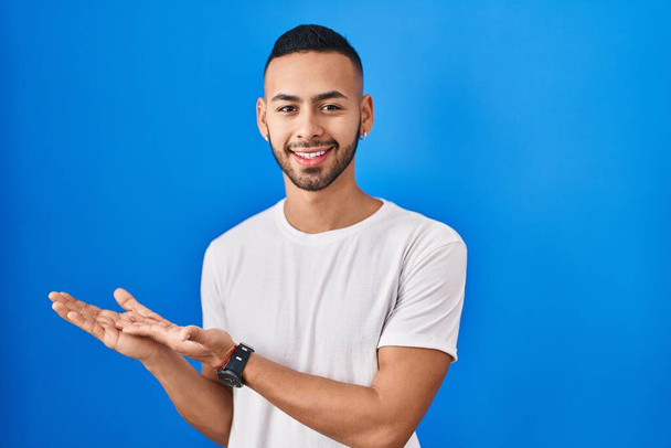 Νεαρός Ισπανόφωνος στέκεται πάνω από το μπλε φόντο δείχνοντας στην άκρη με τα χέρια ανοιχτά παλάμες δείχνει αντίγραφο χώρο, παρουσιάζοντας διαφήμιση χαμογελώντας ενθουσιασμένος χαρούμενος  - Φωτογραφία, εικόνα