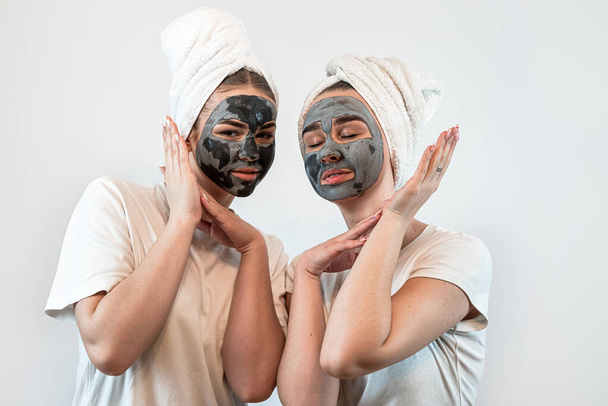 Dos mujeres caucásicas usan toalla con máscara facial de arcilla negra aislada en blanco. Máscara anti edad para hermosa. Concepto de cuidar la piel juvenil - Foto, imagen