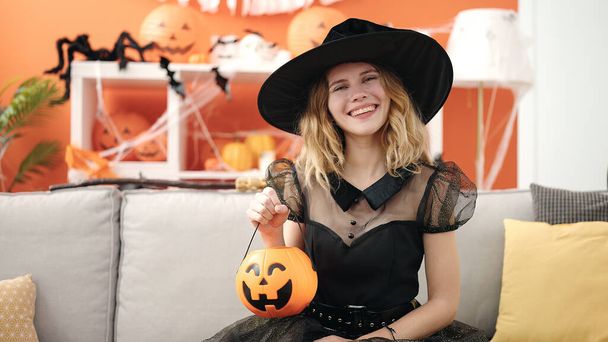Young blonde woman wearing halloween costume holding pumpkin basket at home - 写真・画像