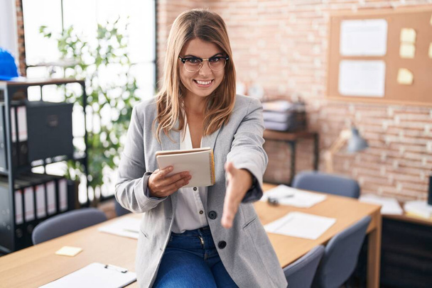 Молода іспаномовна жінка працює в офісі в окулярах, посміхаючись доброзичливо пропонуючи рукостискання як привітання і вітання. успішний бізнес
.  - Фото, зображення