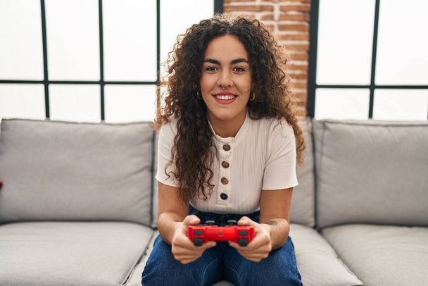 Young beautiful hispanic woman playing video game sitting on sofa at home - 写真・画像