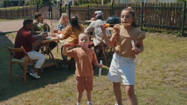Девочка-подросток и её младший брат стоят на заднем дворе и пускают мыльные пузыри на летнюю вечеринку. - Кадры, видео