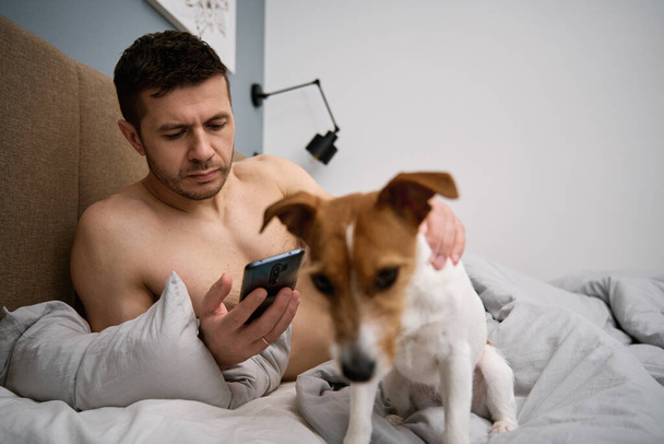 Man met schattige hond chillen in bed 's morgens en gebruik smartphone, luie ochtend. Tijd samen doorbrengen, affectie voor huisdieren - Foto, afbeelding