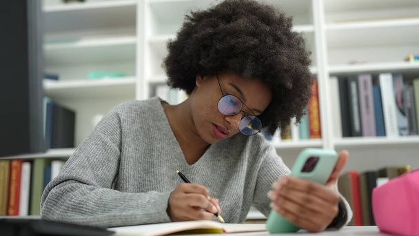 Afrikkalainen amerikkalainen nainen opiskelija, joka käyttää älypuhelimen kirjoittamista muistikirjaan kirjastossa yliopistossa - Valokuva, kuva