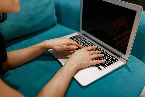 Vágott fotó egy sötét hajú, felnőtt nőről, aki a laptop billentyűzetén gépel, miközben a kanapén fekszik. Online állás- és szabadidős koncepció - Fotó, kép