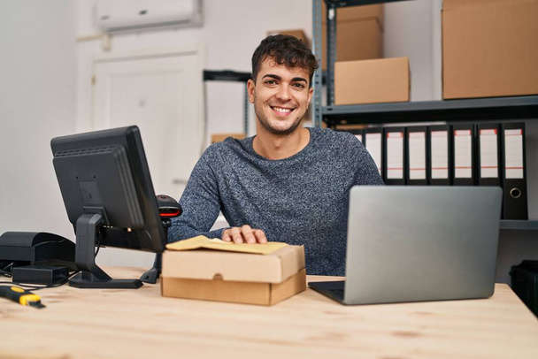 Młody latynoski człowiek e-commerce pracownik za pomocą laptopa trzymając pakiety w biurze - Zdjęcie, obraz