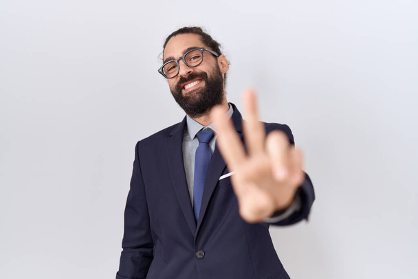 Hombre hispano con barba vistiendo traje y corbata sonriendo mirando a la cámara mostrando los dedos haciendo señal de victoria. número dos.  - Foto, imagen