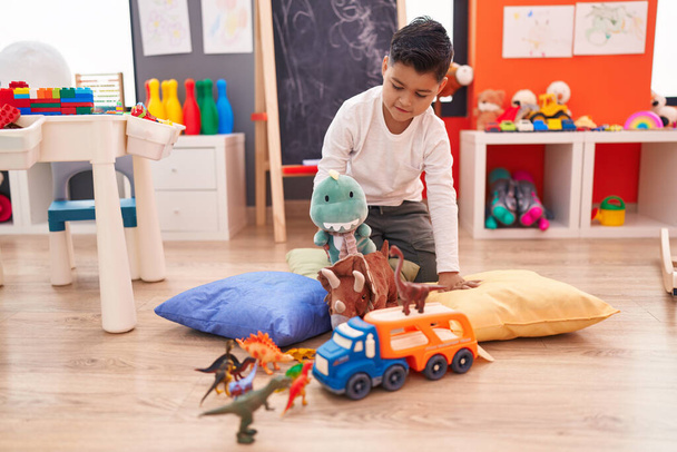 Adorable niño hispano jugando con el coche y el juguete de dinosaurio sentado en el suelo en el jardín de infantes - Foto, Imagen