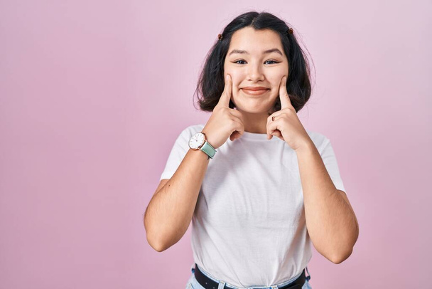Young hispanic woman wearing casual white t shirt over pink background smiling with open mouth, fingers pointing and forcing cheerful smile  - Photo, Image