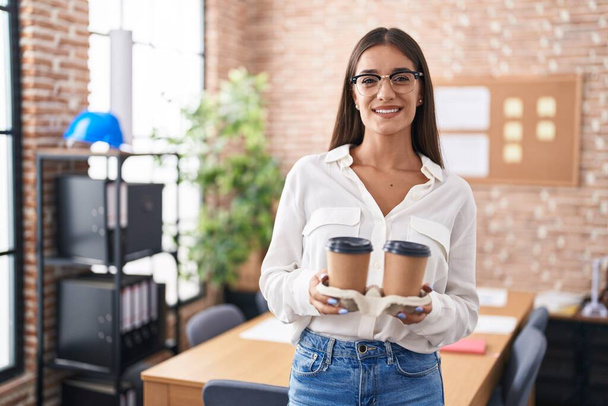 Nuori brunette nainen työskentelee toimistossa tilalla ottaa pois kahvia hymyillen onnellinen ja viileä hymy kasvoilla. hampaiden näyttäminen.  - Valokuva, kuva