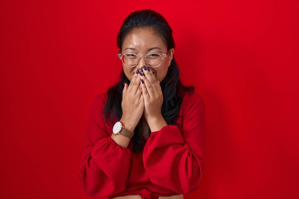 Asian young woman standing over red background laughing and embarrassed giggle covering mouth with hands, gossip and scandal concept  - Foto, immagini