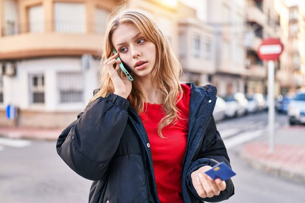 Young blonde woman talking on smartphone holding credit card at street - Foto, Imagem