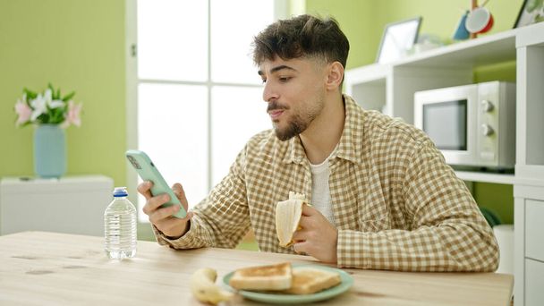 Hombre árabe joven desayunando usando smartphone en casa - Foto, Imagen